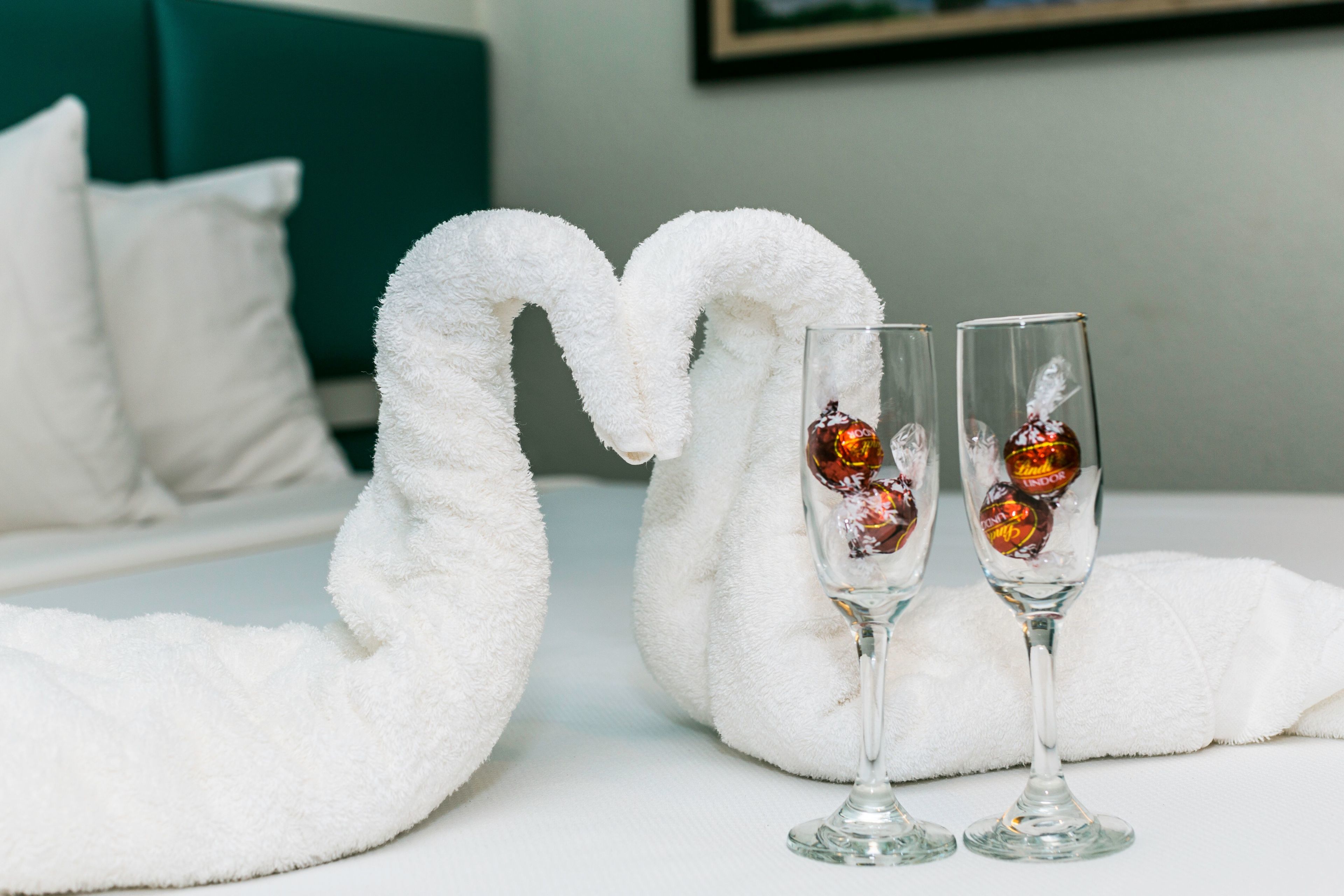 Un lit avec des oreillers blancs et une tête de lit couleur turquoise, deux flûtes à champagne accompagnées de chocolats, et une serviette blanche en forme de cygne.