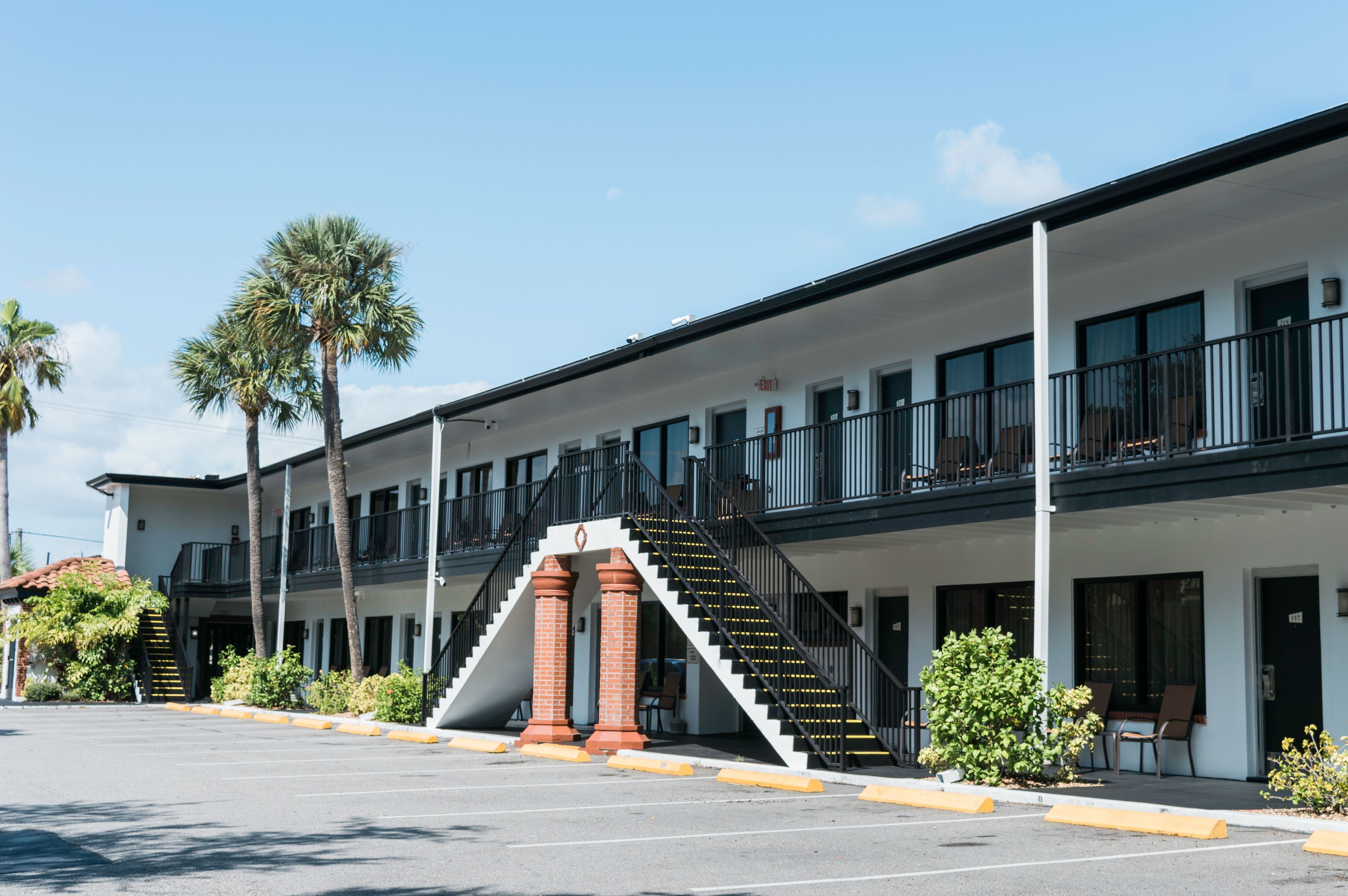 A two-story motel with a central staircase, outdoor seating, and palm trees.