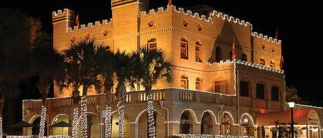Un edificio al estilo castillo con múltiples arcos y un balcón, rodeado de palmeras e iluminado con luces.