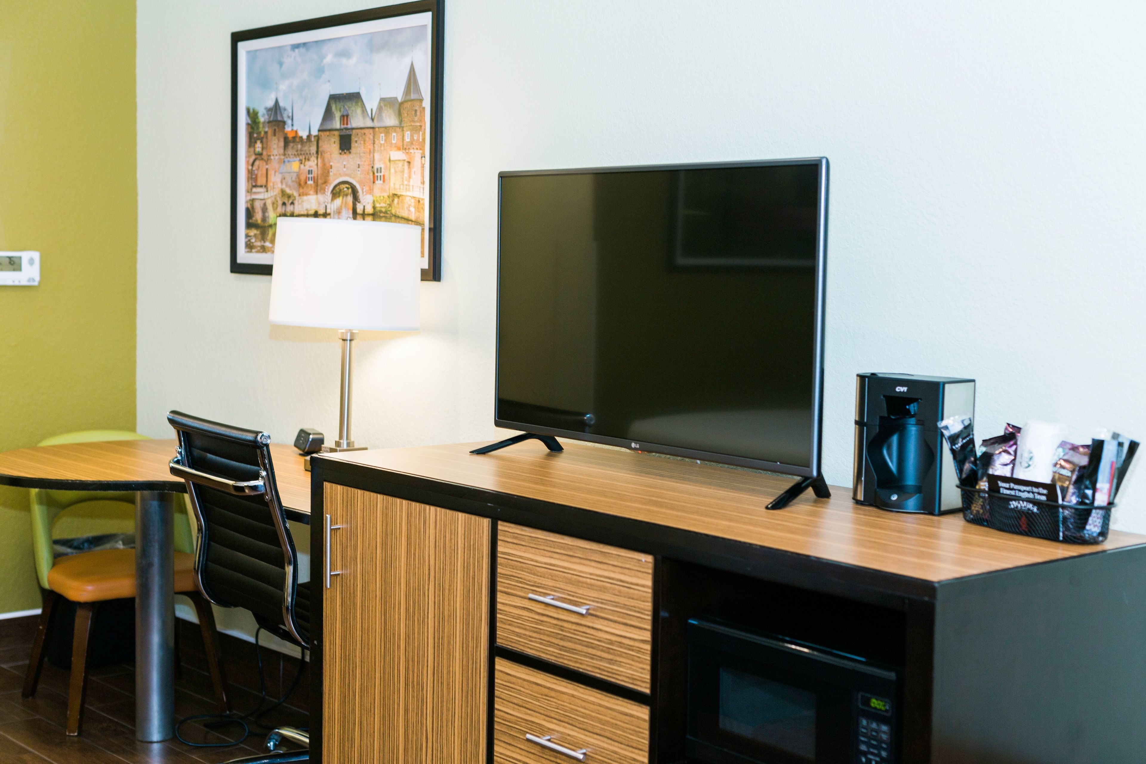 A hotel room with a wooden desk, a flat-screen TV, a lamp, a coffee maker, and a framed picture on the wall.