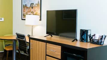 A hotel room with a wooden desk, a flat-screen TV, a lamp, a coffee maker, and a framed picture on the wall.