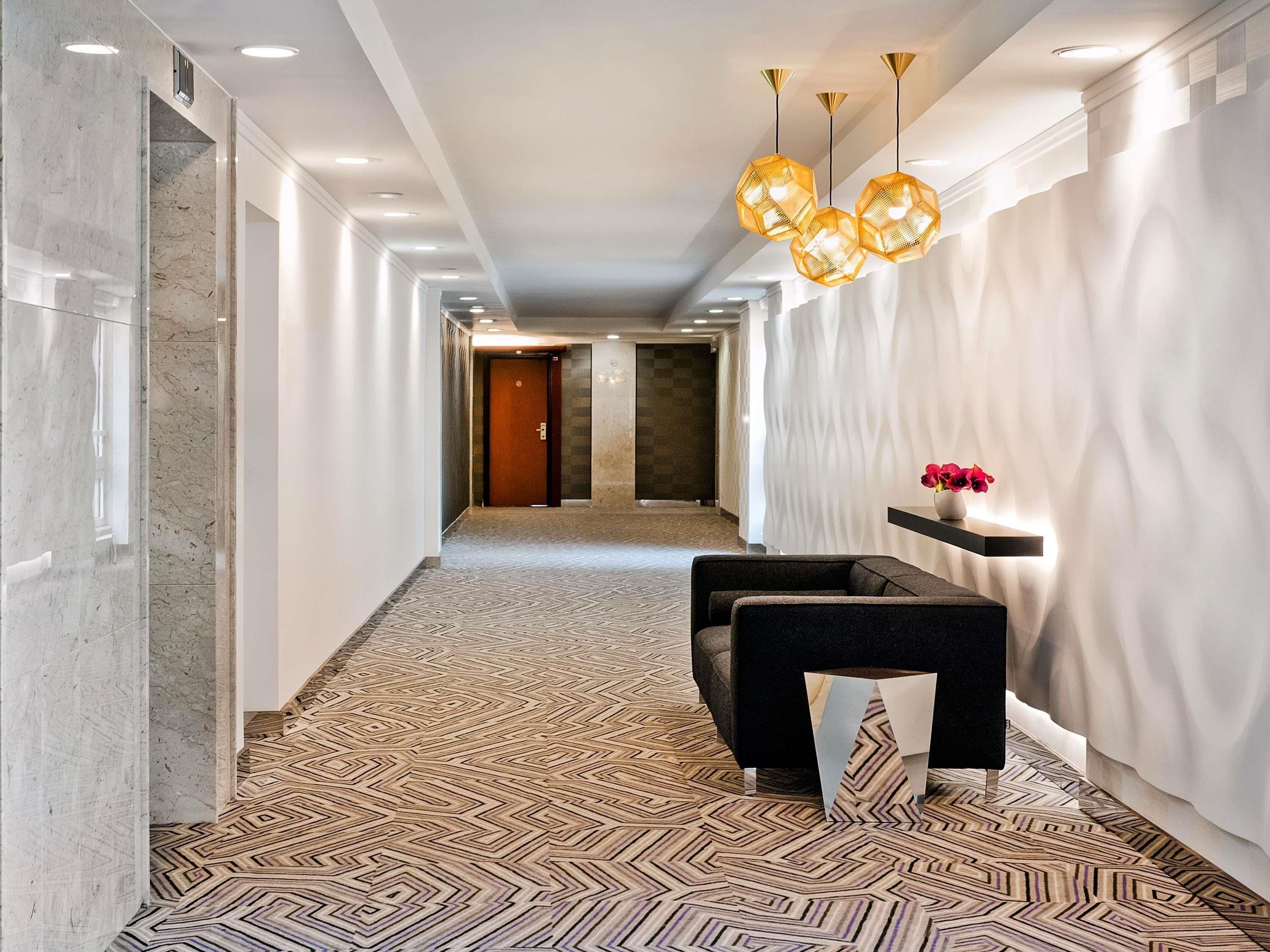 A hallway with a patterned carpet, a reception desk, a wall-mounted shelf with flowers, and a door at the end.