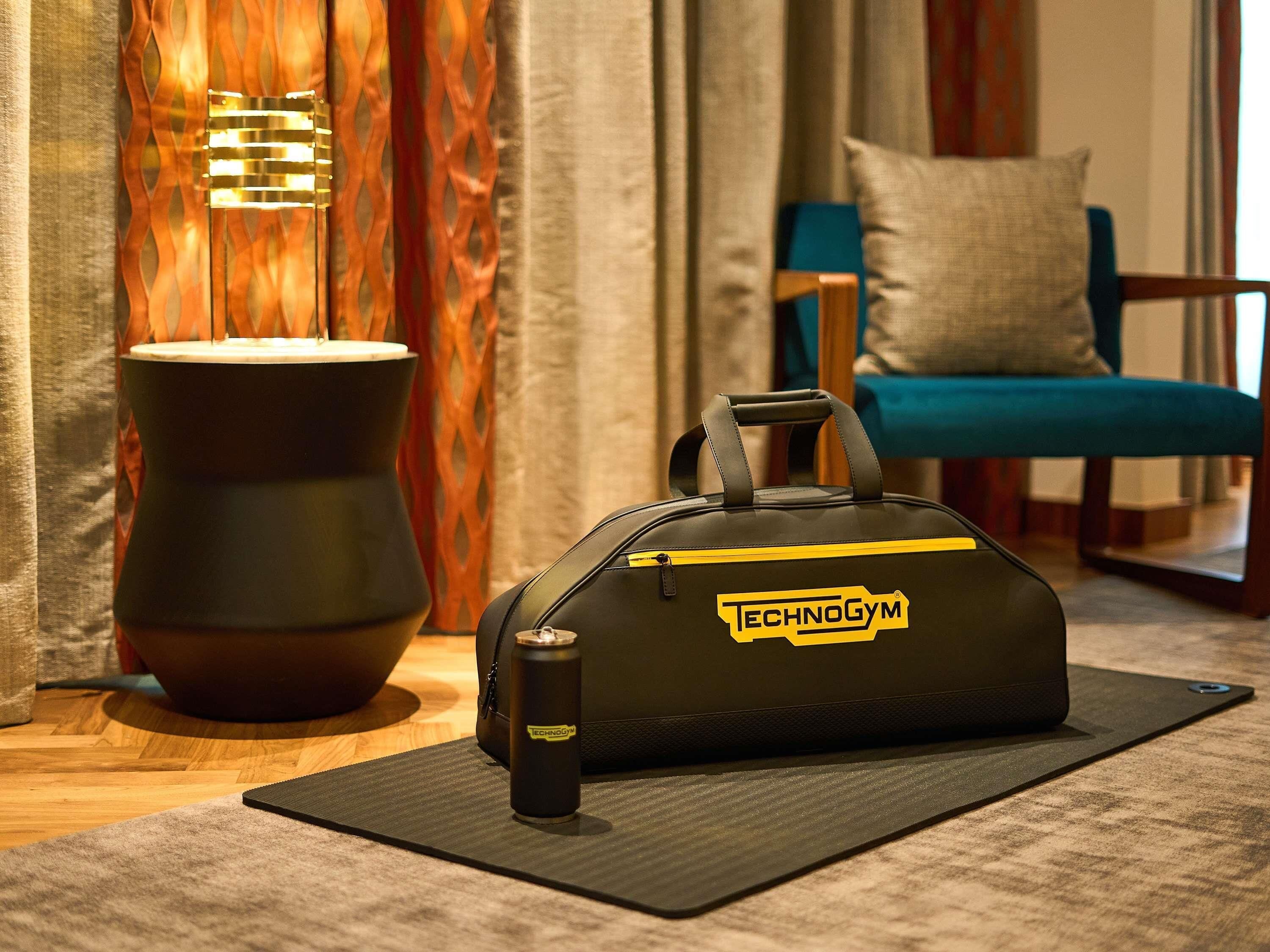 Una habitación con una bolsa negra que dice 'TECHNOGYM', un pequeño recipiente, una colchoneta negra, una silla azul y un jarrón decorativo.