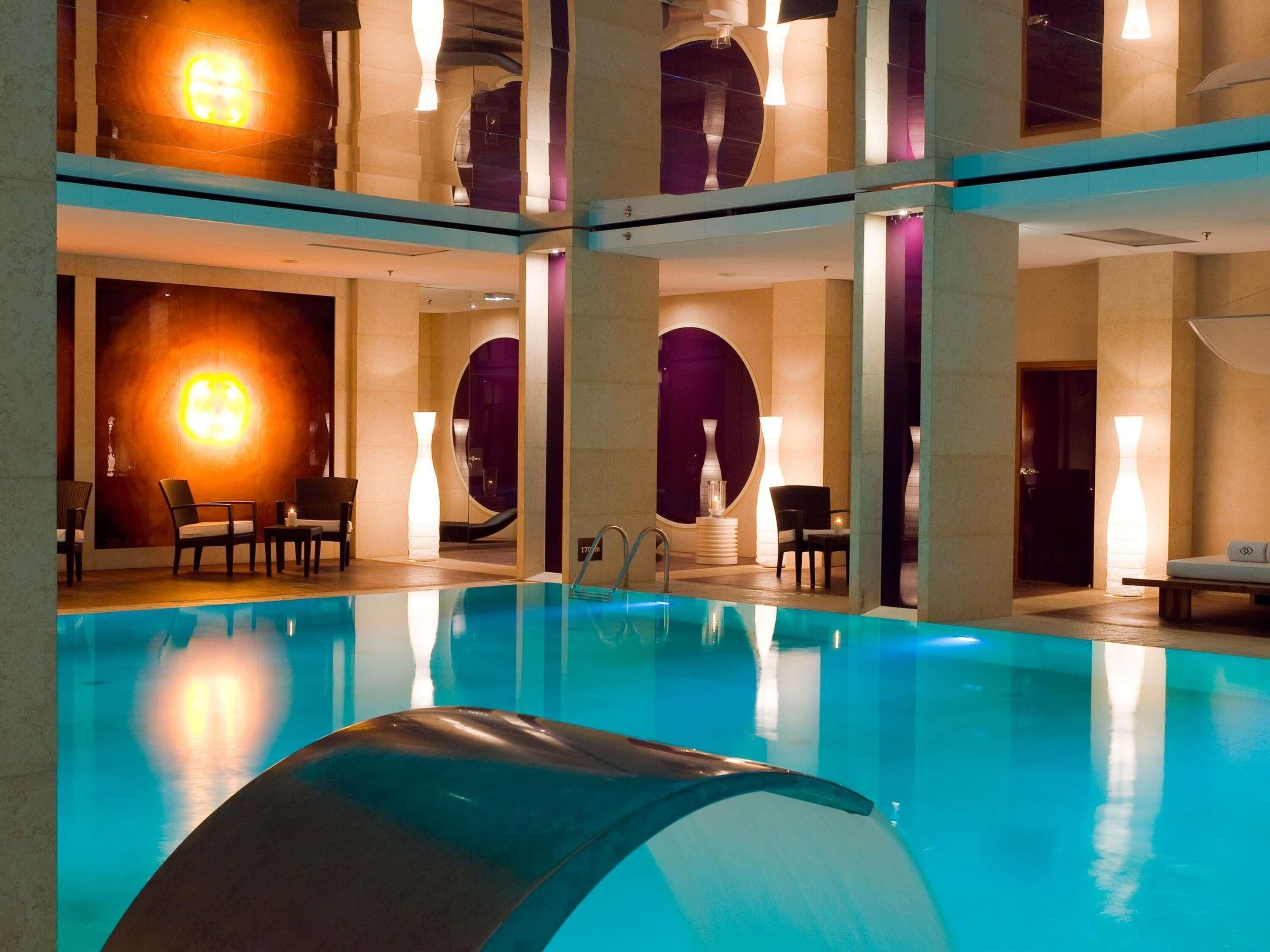 A multi-level indoor swimming pool with clear water, lounge chairs, and modern lighting fixtures.
