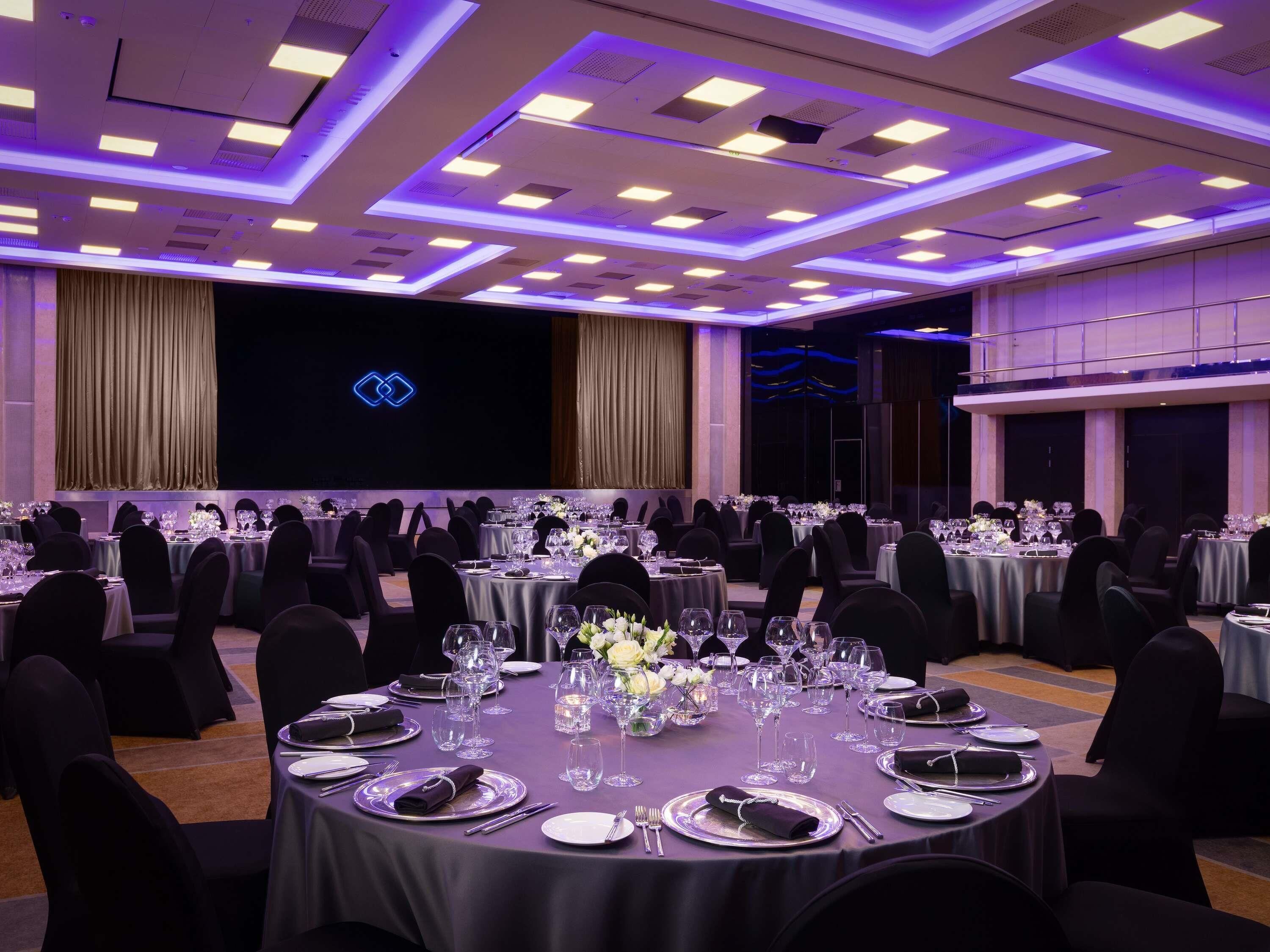 Un salón de banquetes con mesas redondas dispuestas para el servicio de comidas, un escenario con un logotipo azul y un techo alto con iluminación empotrada.