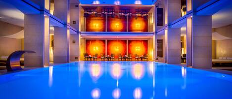 Una piscina interior moderna con suelo azul, rodeada de zonas de estar y una pared cálida e iluminada.