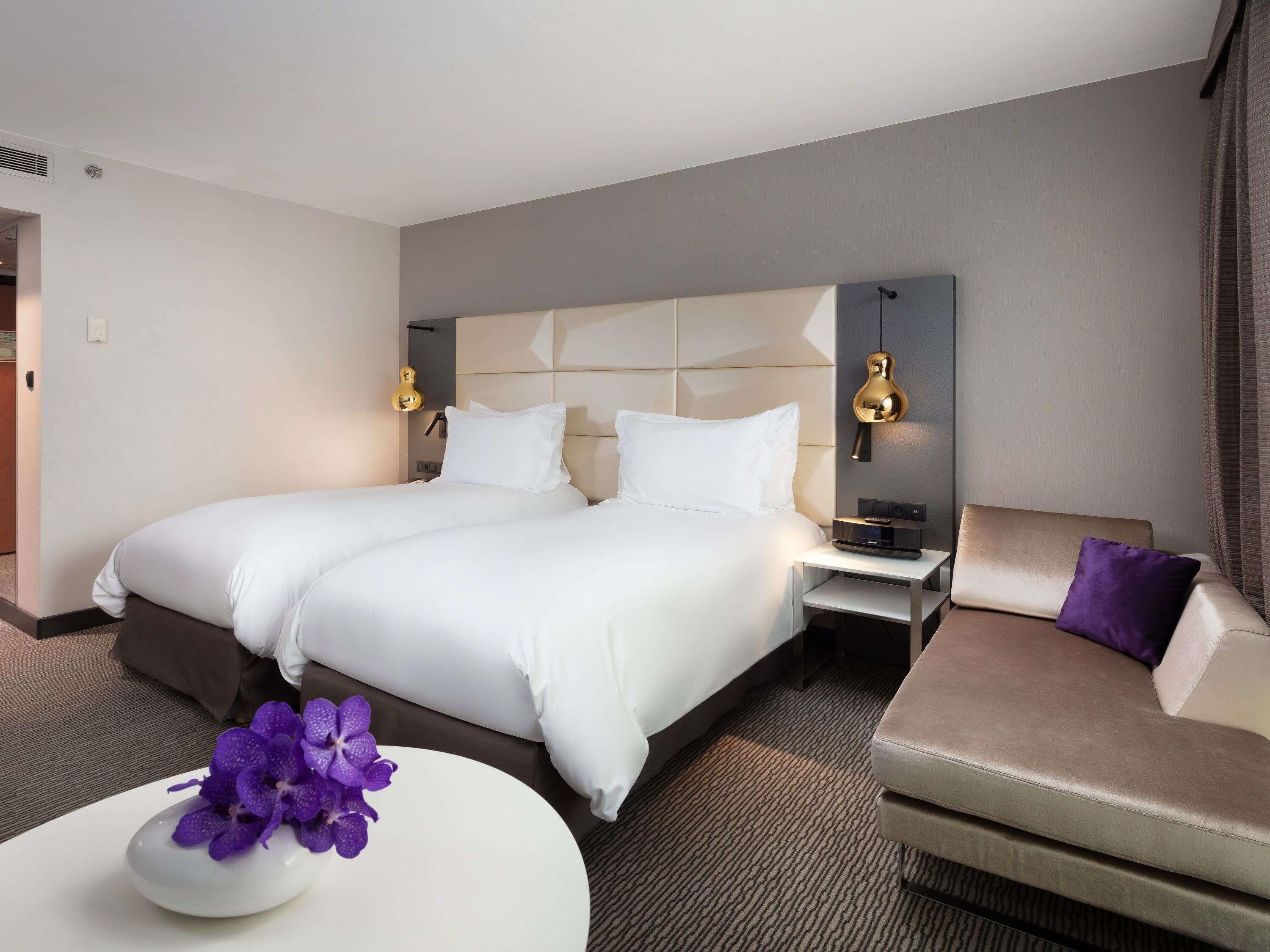Une chambre d’hôtel avec un grand lit, deux tables de chevet, un canapé et une petite table sur laquelle est posée une orchidée violette.
