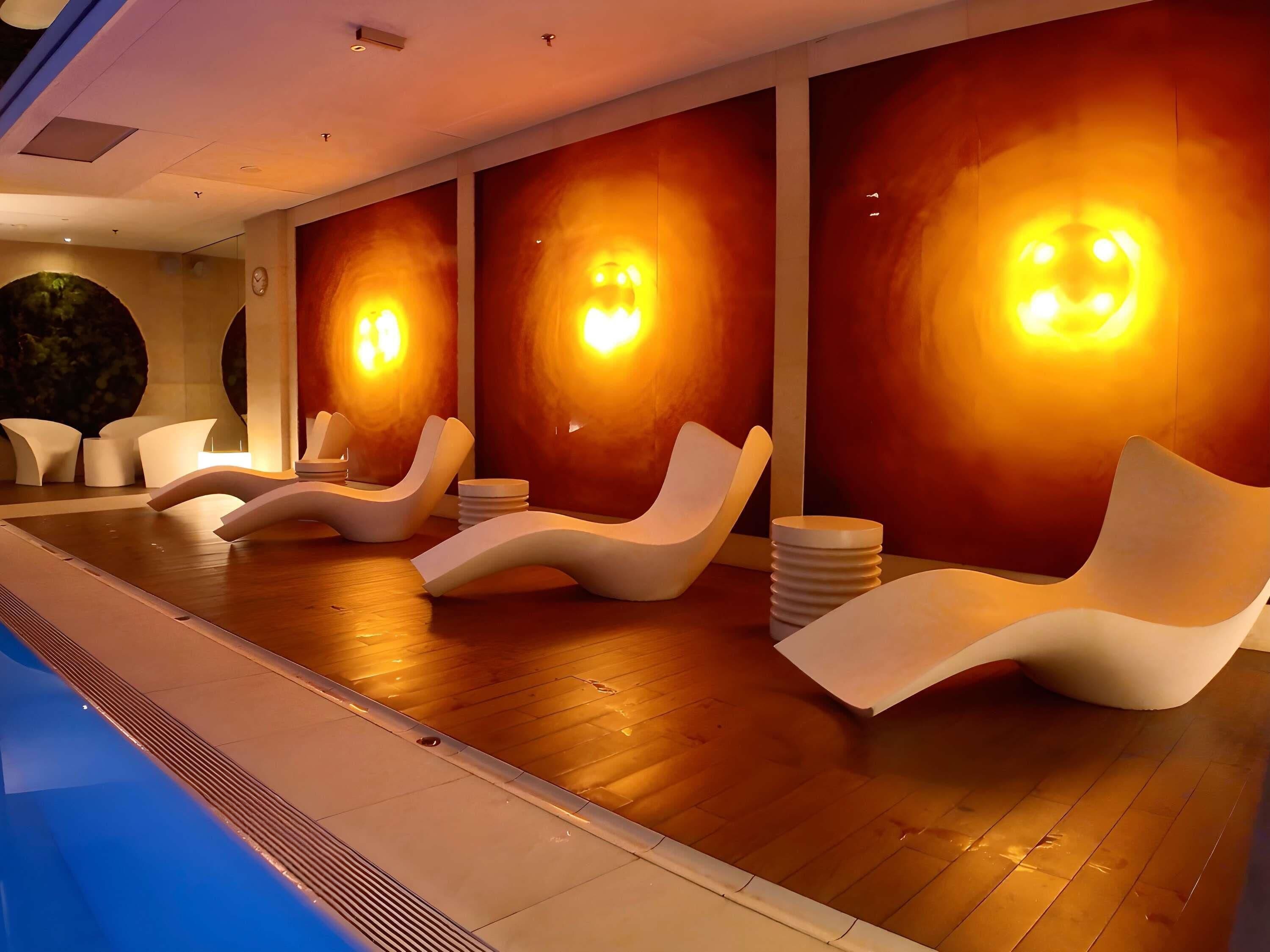 A modern indoor pool area with lounge chairs and wall-mounted lighting.