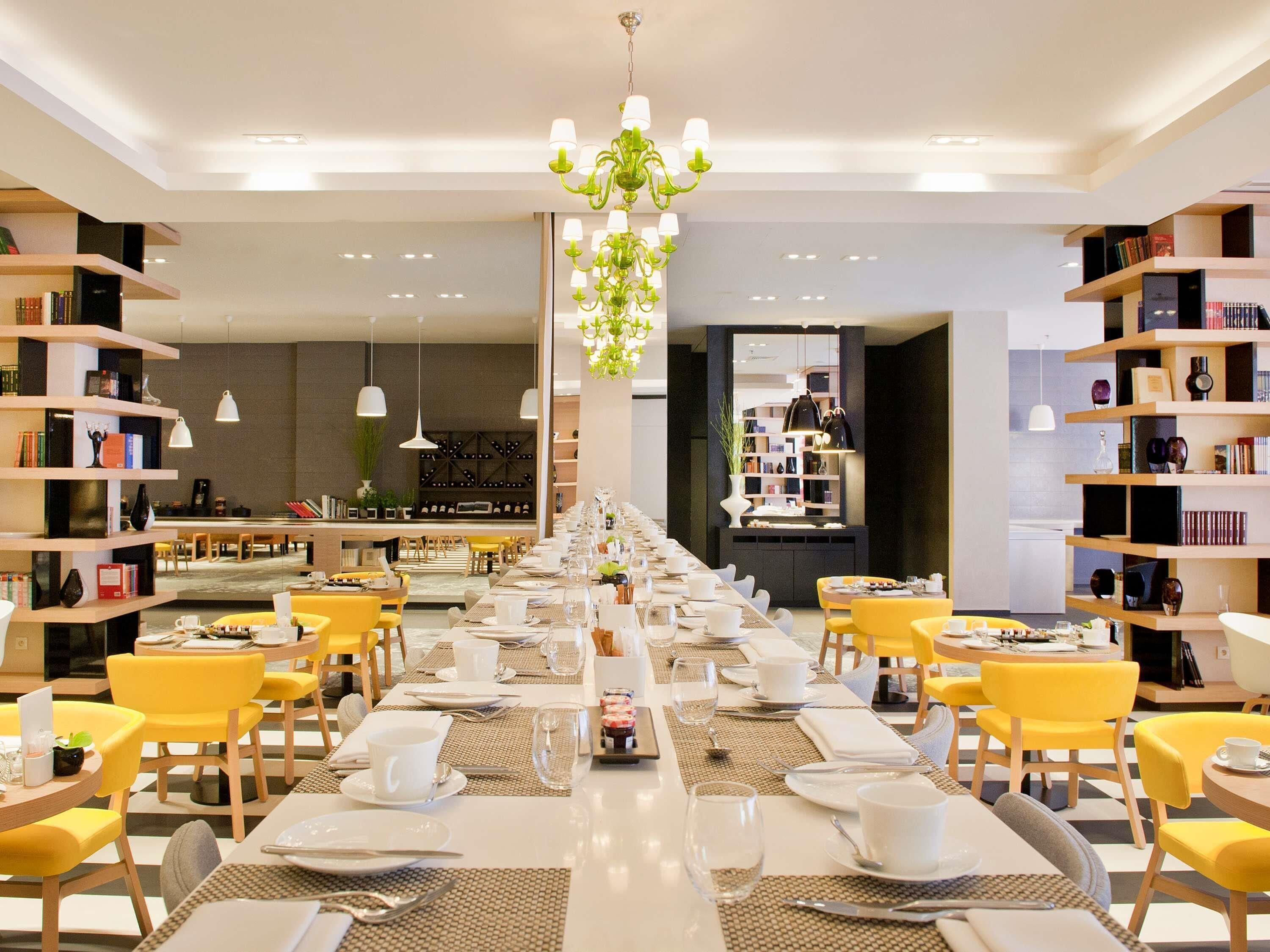 Un comedor de hotel con mesas largas, sillas amarillas y estanterías.