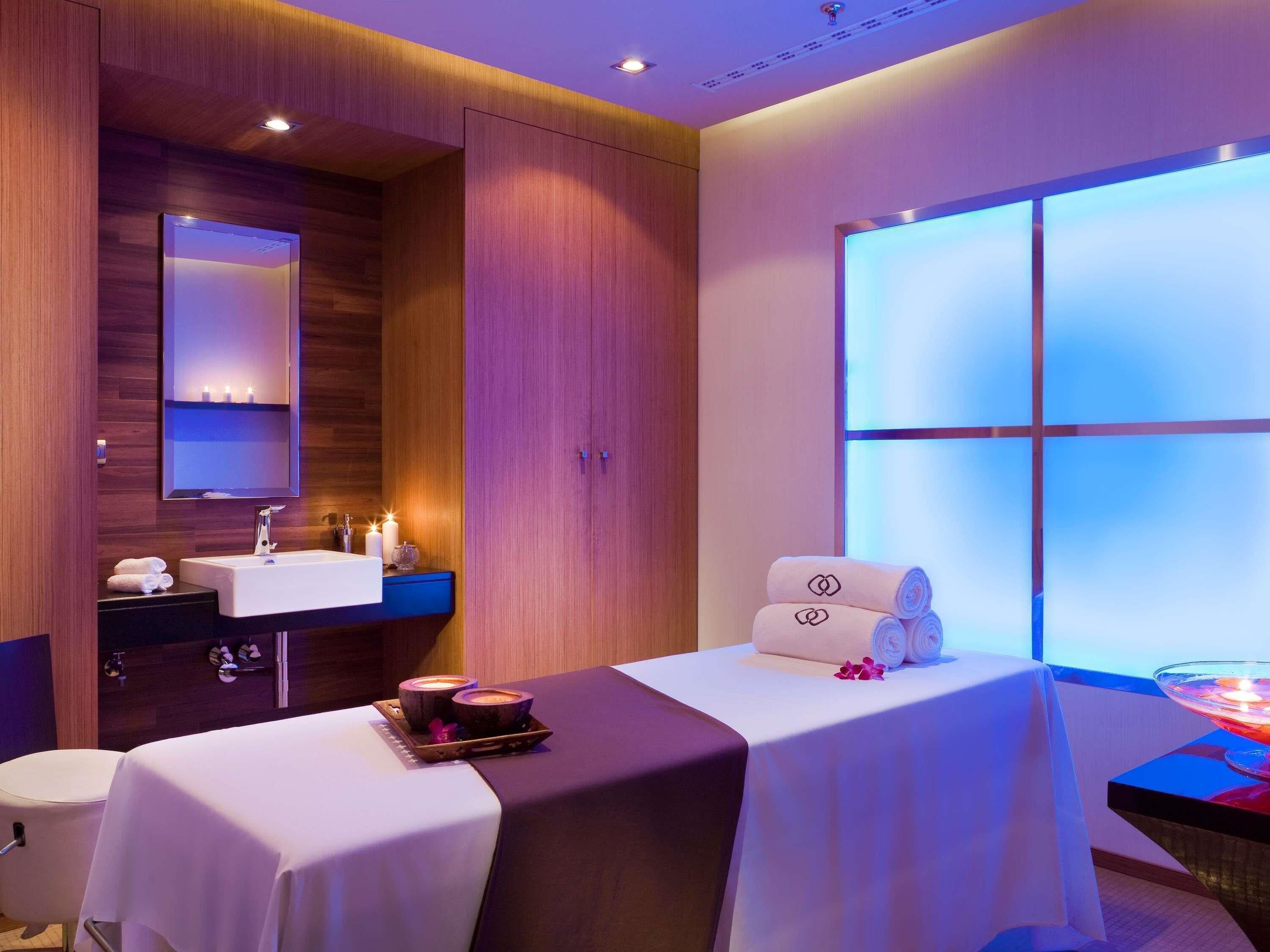 A modern hotel room with a wooden wall, a large mirror, a white sink, a massage table, and a large window with a view.