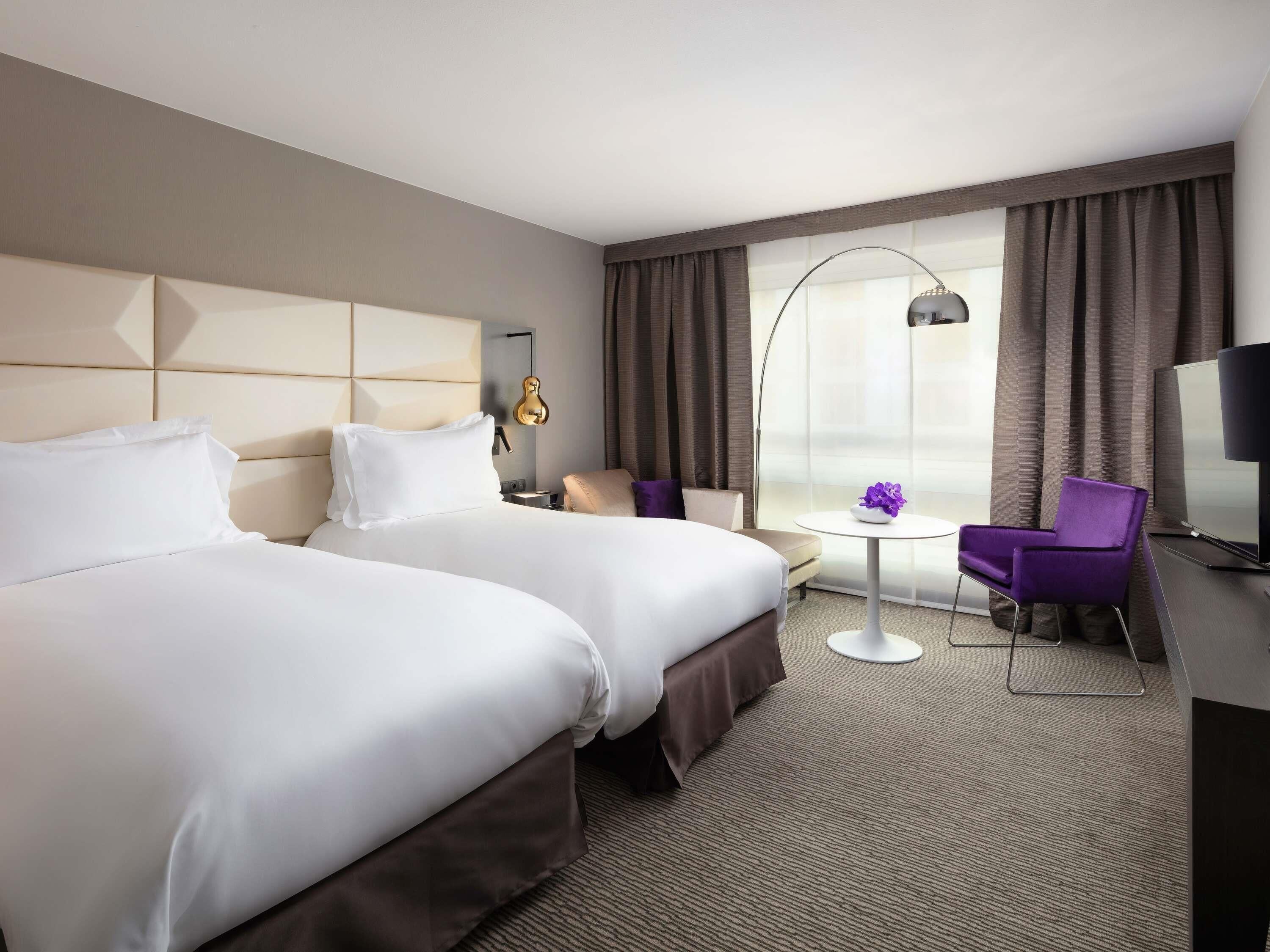 A hotel room with two beds, a desk, a chair, a TV, and a window with curtains.