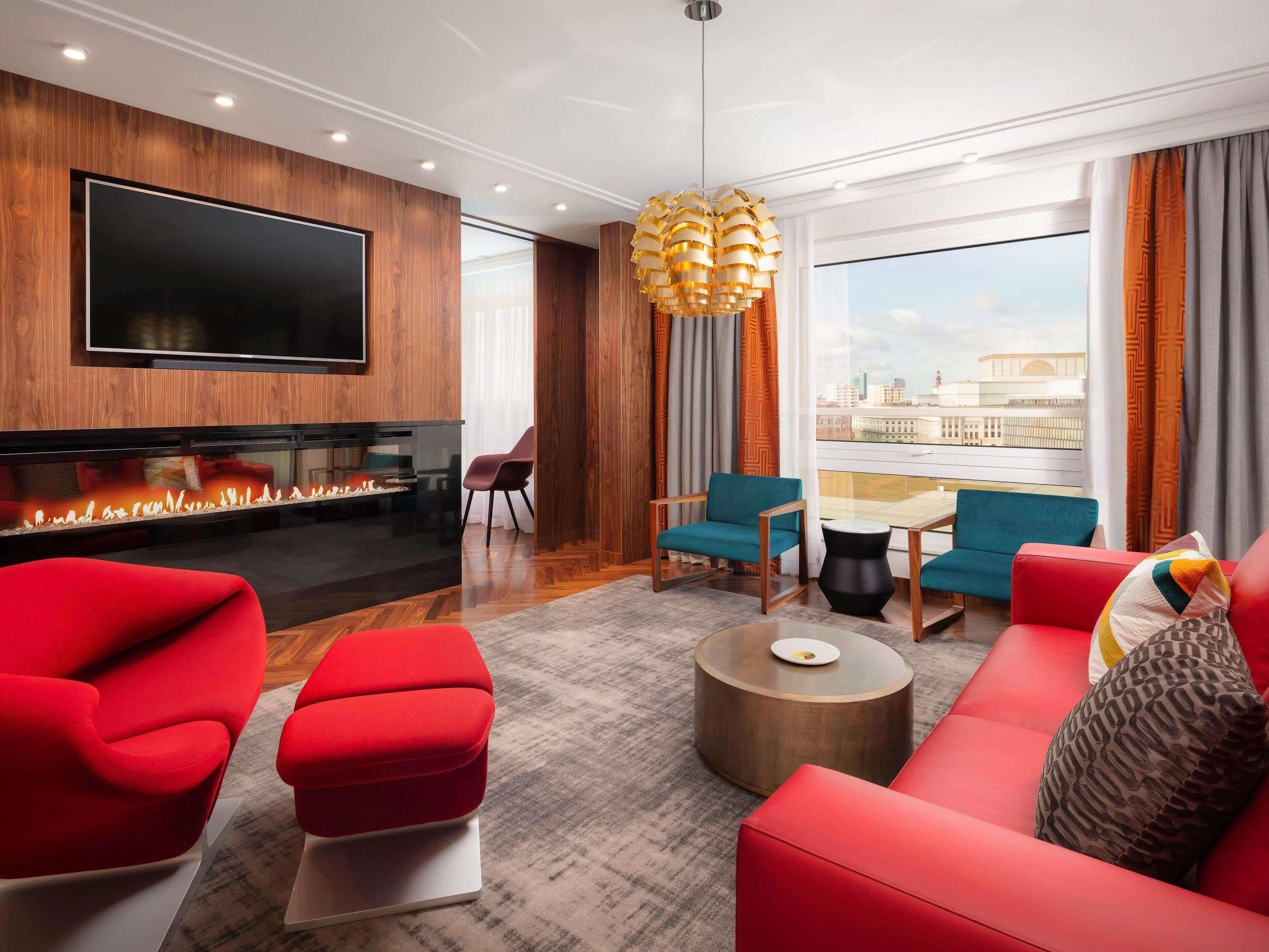 A modern living room with a flat-screen TV, a fireplace, red and blue seating, and a large window with a city view.