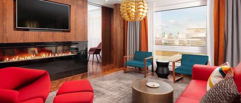 A modern living room with a flat-screen TV, a fireplace, red and blue seating, and a large window with a city view.