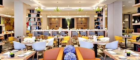 A modern dining area with multiple tables and chairs, a central decorative arrangement, and bookshelves on the sides.
