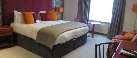 A hotel room with a large bed, a small table, a chair, and a window with curtains.