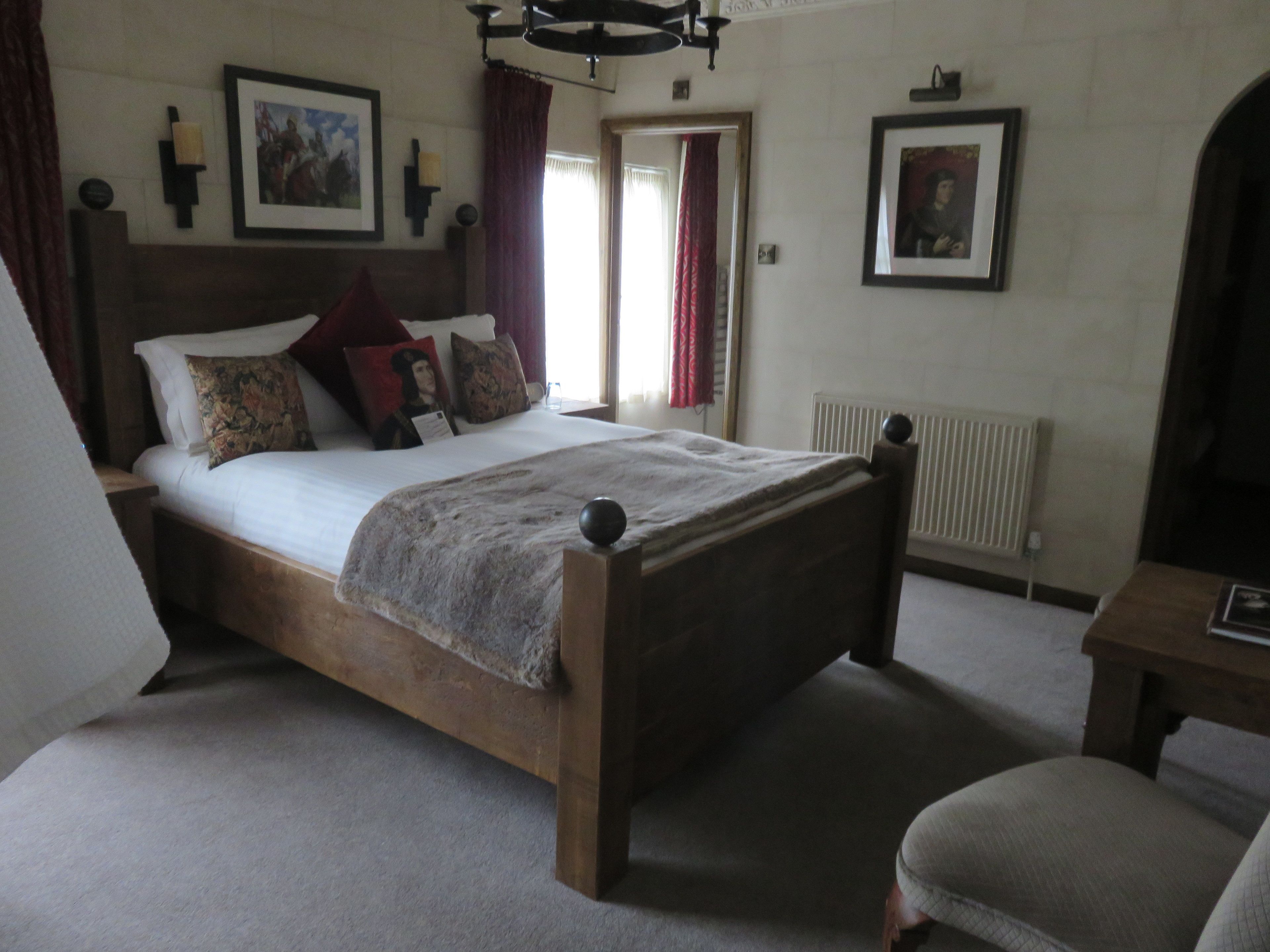 Una cama de madera tradicional con cabecera, una mesita de noche y una silla. Hay dos cuadros enmarcados en la pared, un radiador y una ventana con cortinas.