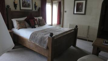Una cama de madera tradicional con cabecera, una mesita de noche y una silla. Hay dos cuadros enmarcados en la pared, un radiador y una ventana con cortinas.