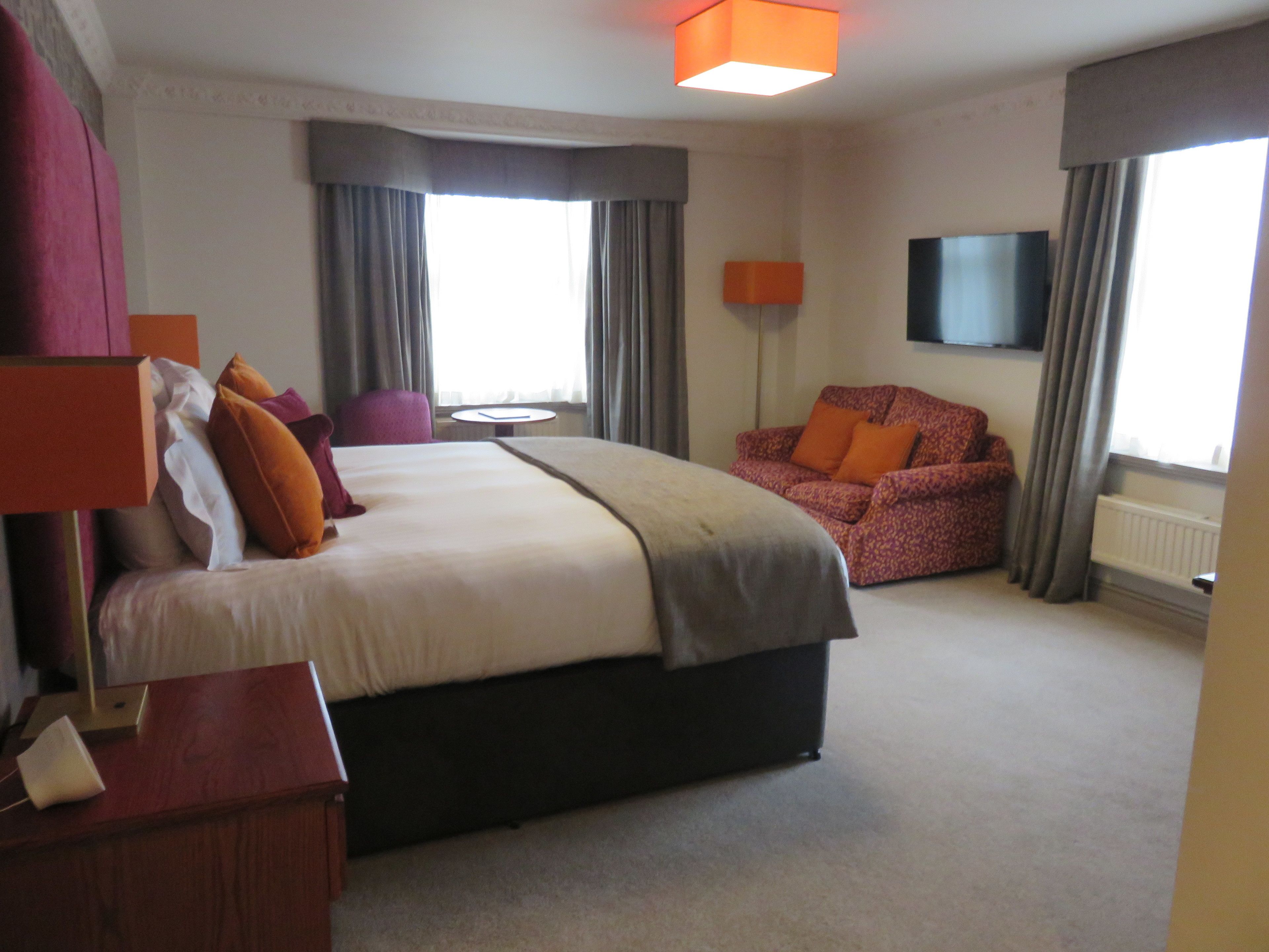 A hotel room with a large bed, a desk, a chair, a sofa, and a TV on the wall.