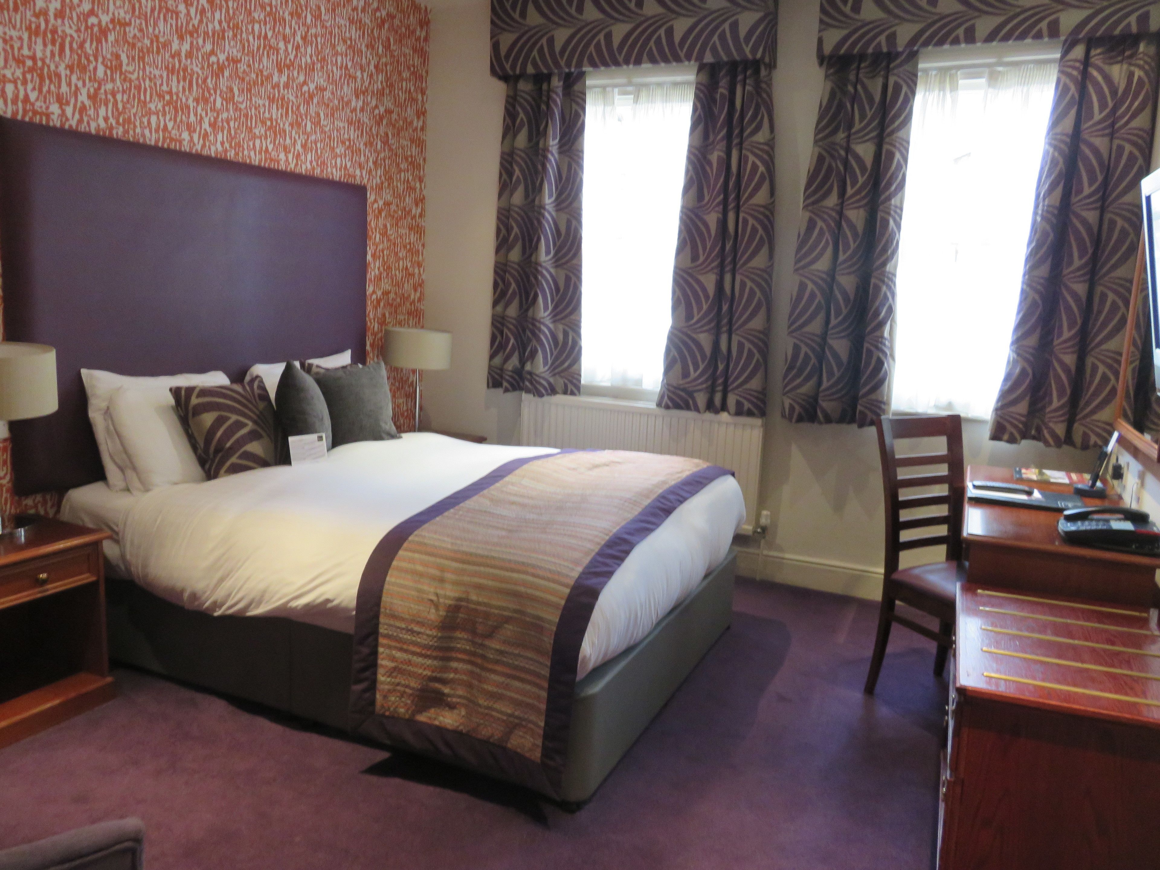 A hotel room with a large bed, a desk, and a chair. There are two windows with curtains, a radiator, and a wall with a patterned wallpaper.