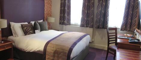 A hotel room with a large bed, a desk, and a chair. There are two windows with curtains, a radiator, and a wall with a patterned wallpaper.