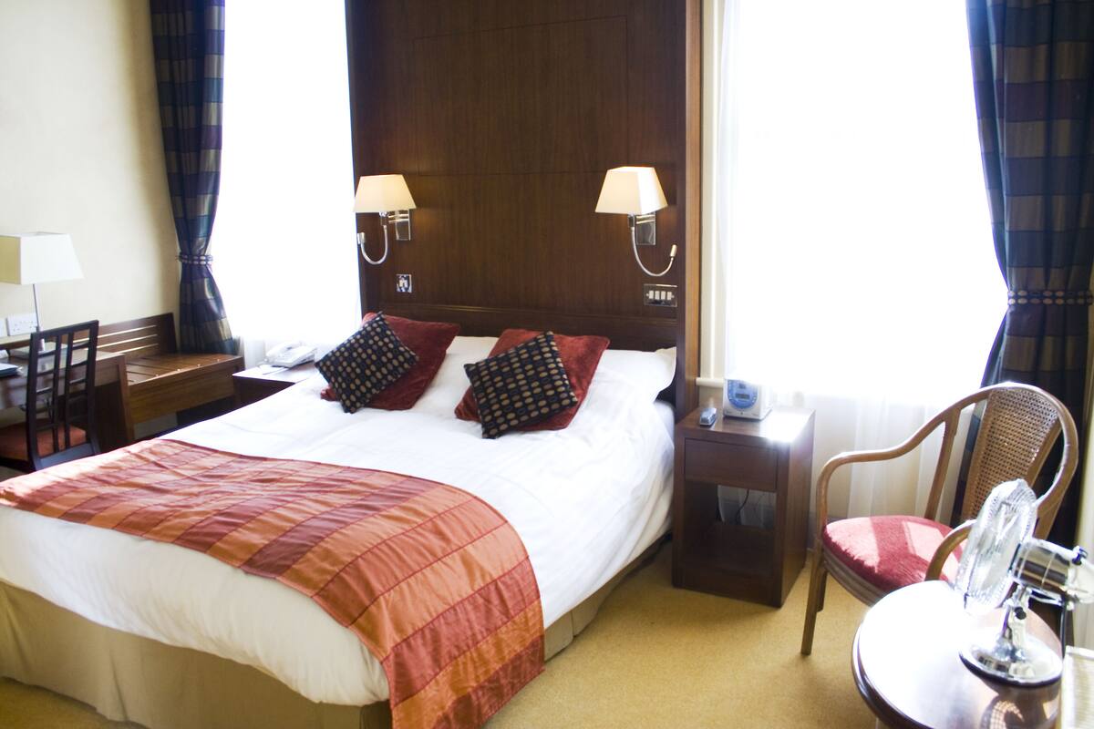 A hotel room with a large bed, two bedside lamps, a wooden headboard, a desk with a chair, a chair with a red cushion, and a small table with a mirror.
