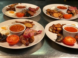 Una mesa de desayuno con platos de huevos, tocino, salchichas, champiñones y tomates, acompañados de recipientes pequeños de salsa de tomate.