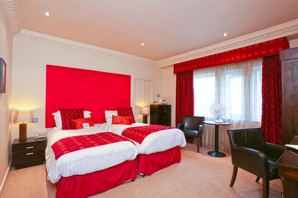 A hotel room with two beds, a desk, a chair, and a small table with a fan.