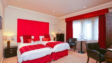 A hotel room with two beds, a desk, a chair, and a small table with a fan.