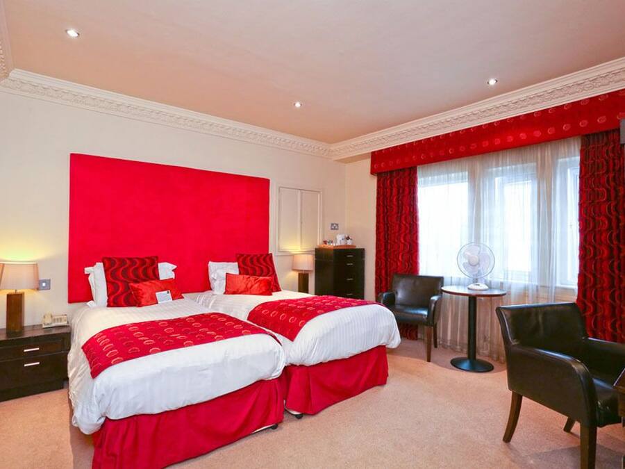 A hotel room with two beds, a desk, a chair, and a small table with a fan.