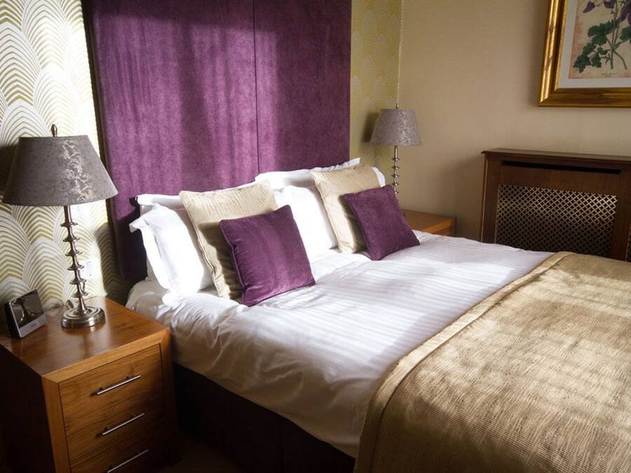 Una cama bien hecha con ropa de cama blanca y morada, una mesita de noche de madera con una lámpara e impresión botánica enmarcada en la pared.