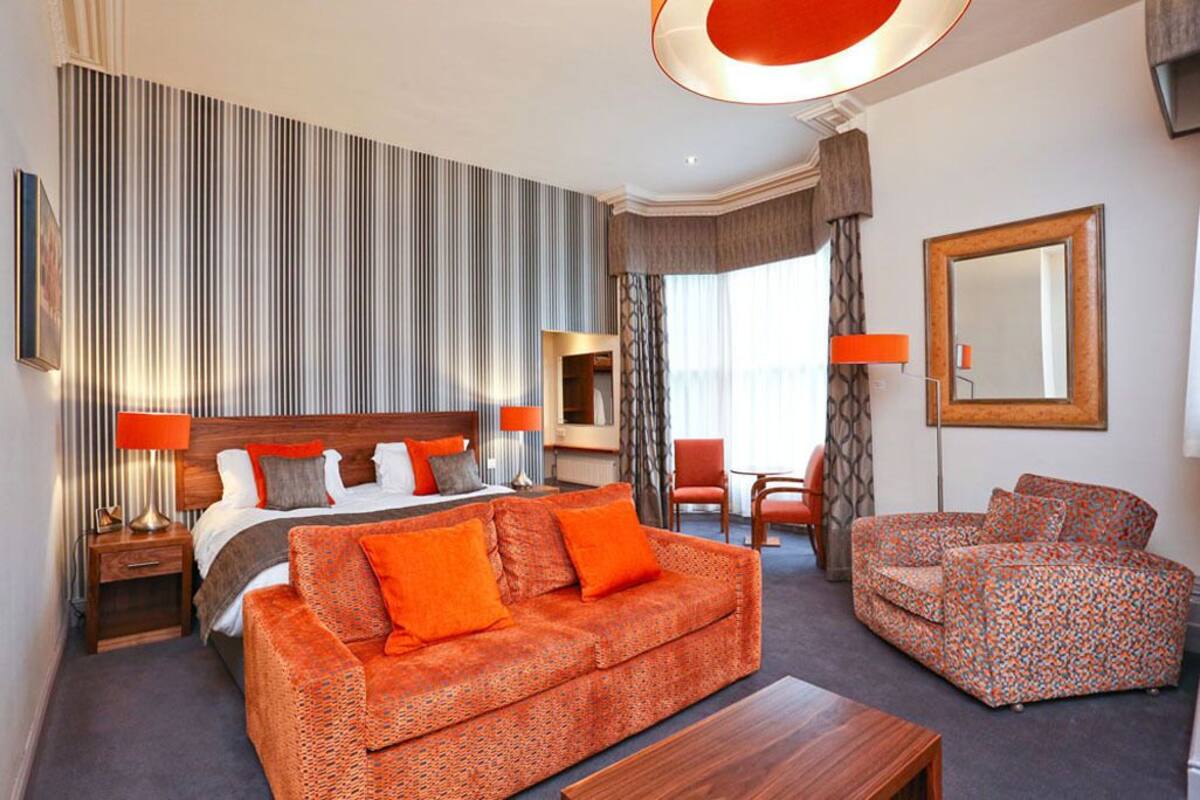 A hotel room with a bed, orange patterned sofa, a wooden coffee table, a chair, a mirror, and a striped wall.