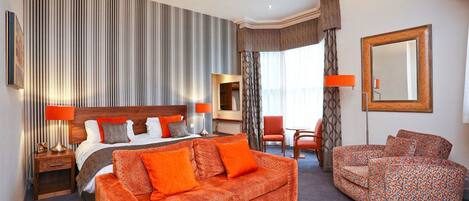 A hotel room with a bed, orange patterned sofa, a wooden coffee table, a chair, a mirror, and a striped wall.