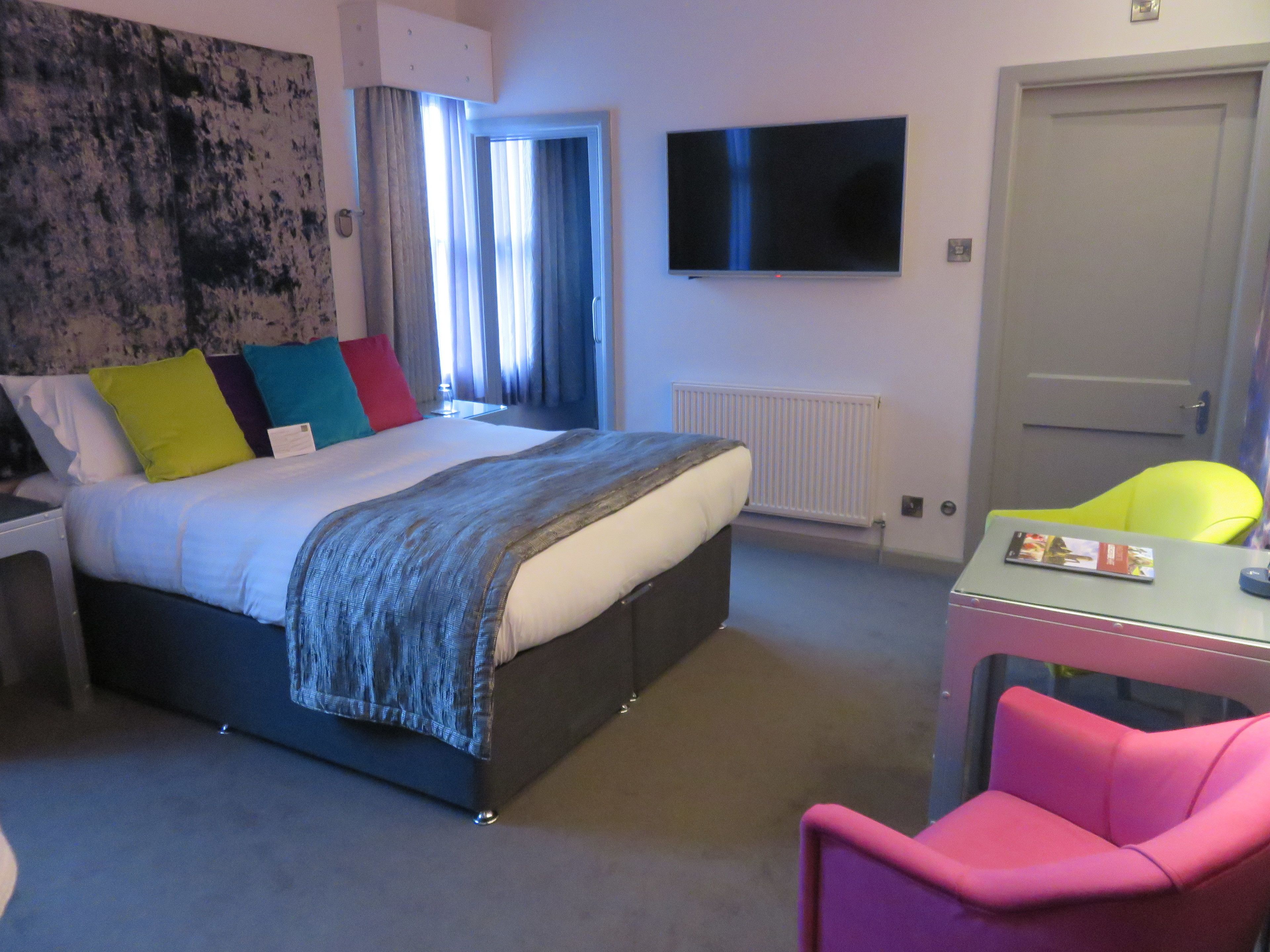 A hotel room with a bed, a TV mounted on the wall, a desk with a chair, and a door.