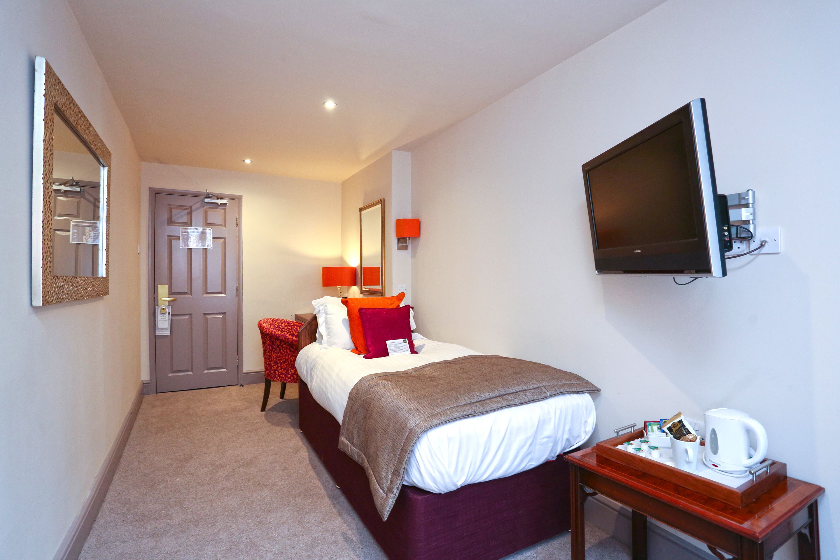 A hotel room with a bed, a TV mounted on the wall, a bedside table with a coffee maker, and a door with a mirror and light fixture.