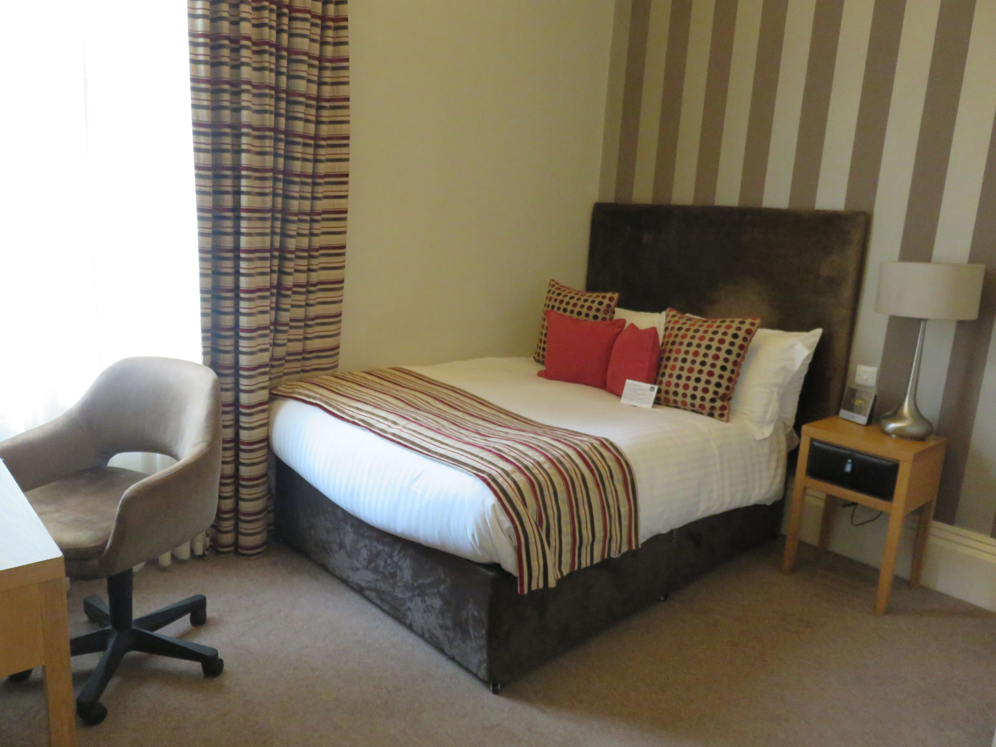 A hotel room with a bed, a desk, a chair, a nightstand, and a lamp.