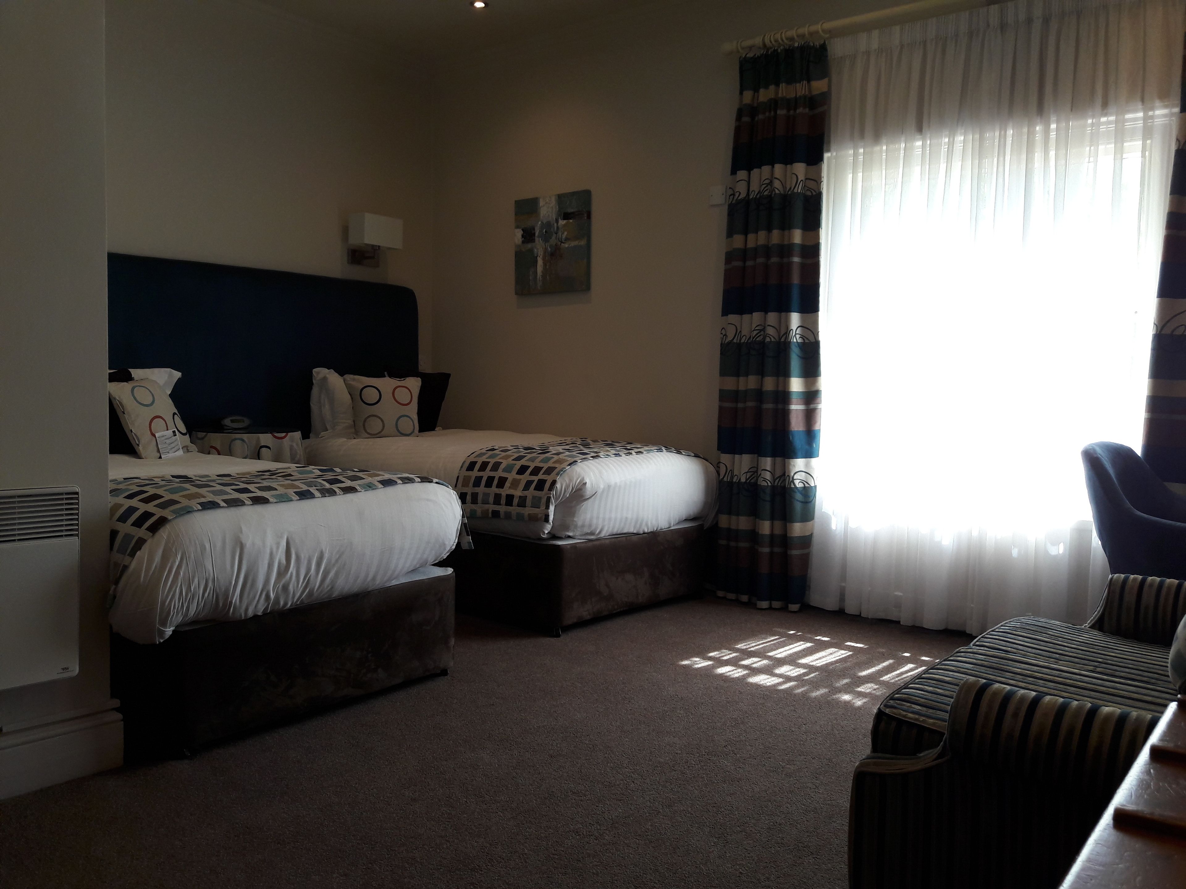 A hotel room with two beds, a chair, a desk, a radiator, and a window with curtains.