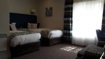 A hotel room with two beds, a chair, a desk, a radiator, and a window with curtains.
