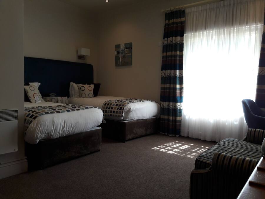 A hotel room with two beds, a chair, a desk, a radiator, and a window with curtains.