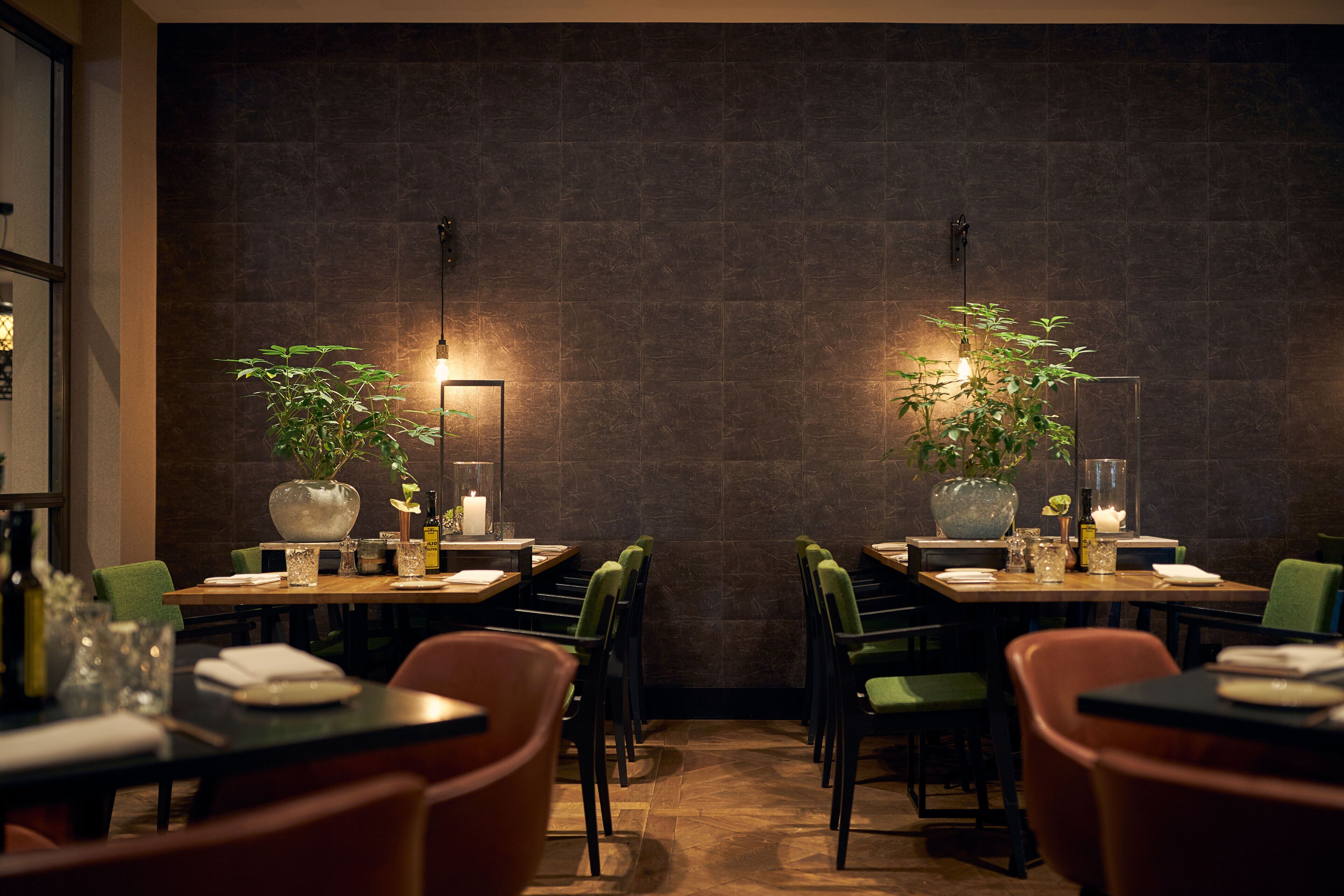 Een restaurant met donkere betegelde muren, groene stoelen en tafels gedekt met kaarsen en planten.