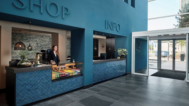 A modern hotel lobby with a blue reception desk labeled "SHOP" and "INFO," featuring a fruit display and a receptionist. - Van der Valk Hotel Antwerpen (Antwerp)