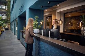 A reception desk with a modern design, a potted plant, and a large window with a view. - Van der Valk Hotel Antwerpen (Antwerp)