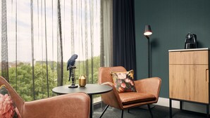 A room with a view of greenery through sheer curtains, a round table with a lamp, a sofa with a floral pillow, and a wooden cabinet. - Van der Valk Hotel Antwerpen (Antwerp)