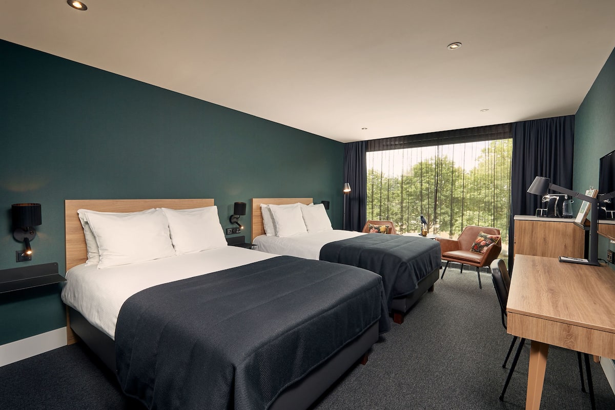 A hotel room with two beds, a desk, a chair, and a large window with a view of trees.