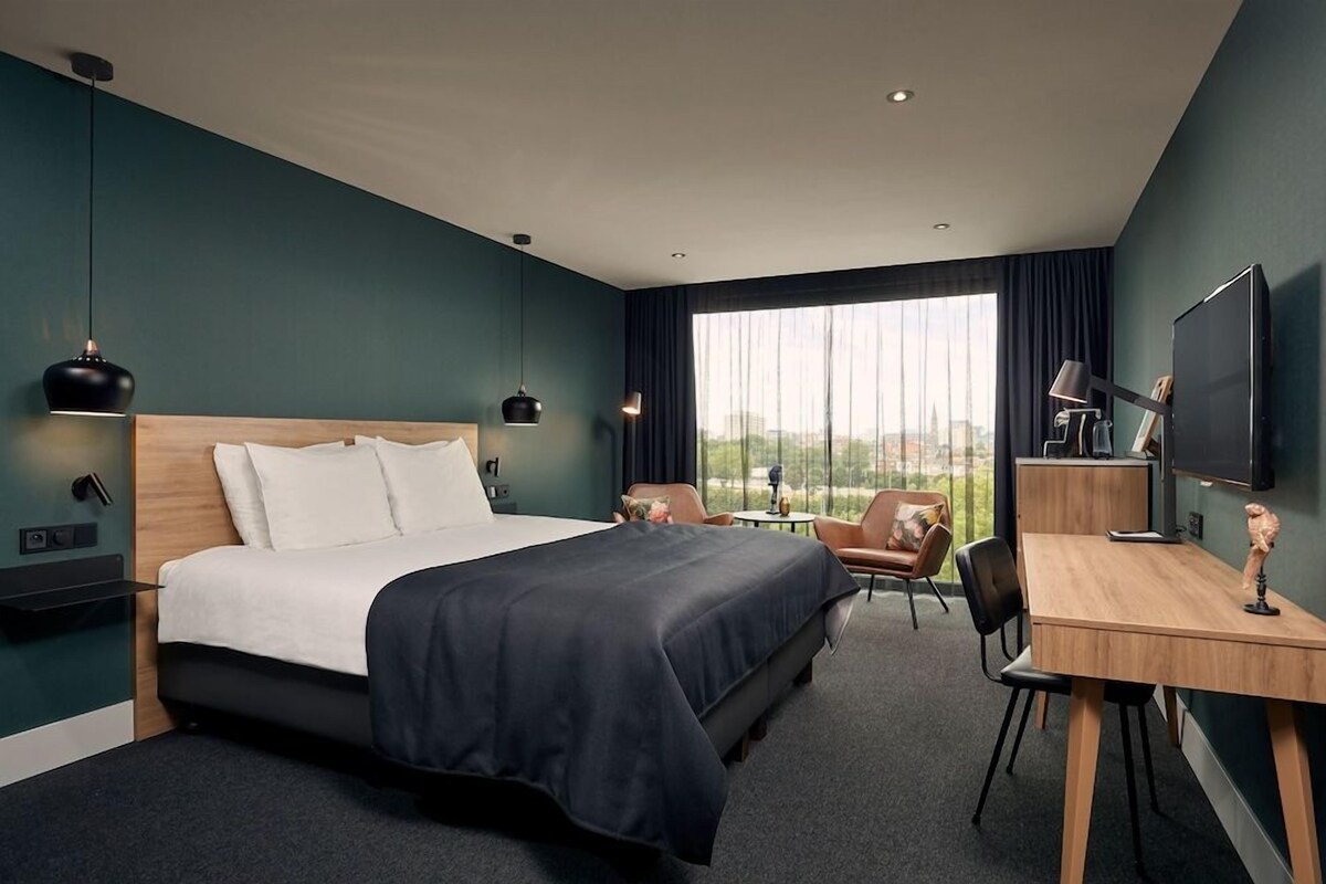 A modern hotel room with a large bed, a desk, a chair, and a TV.