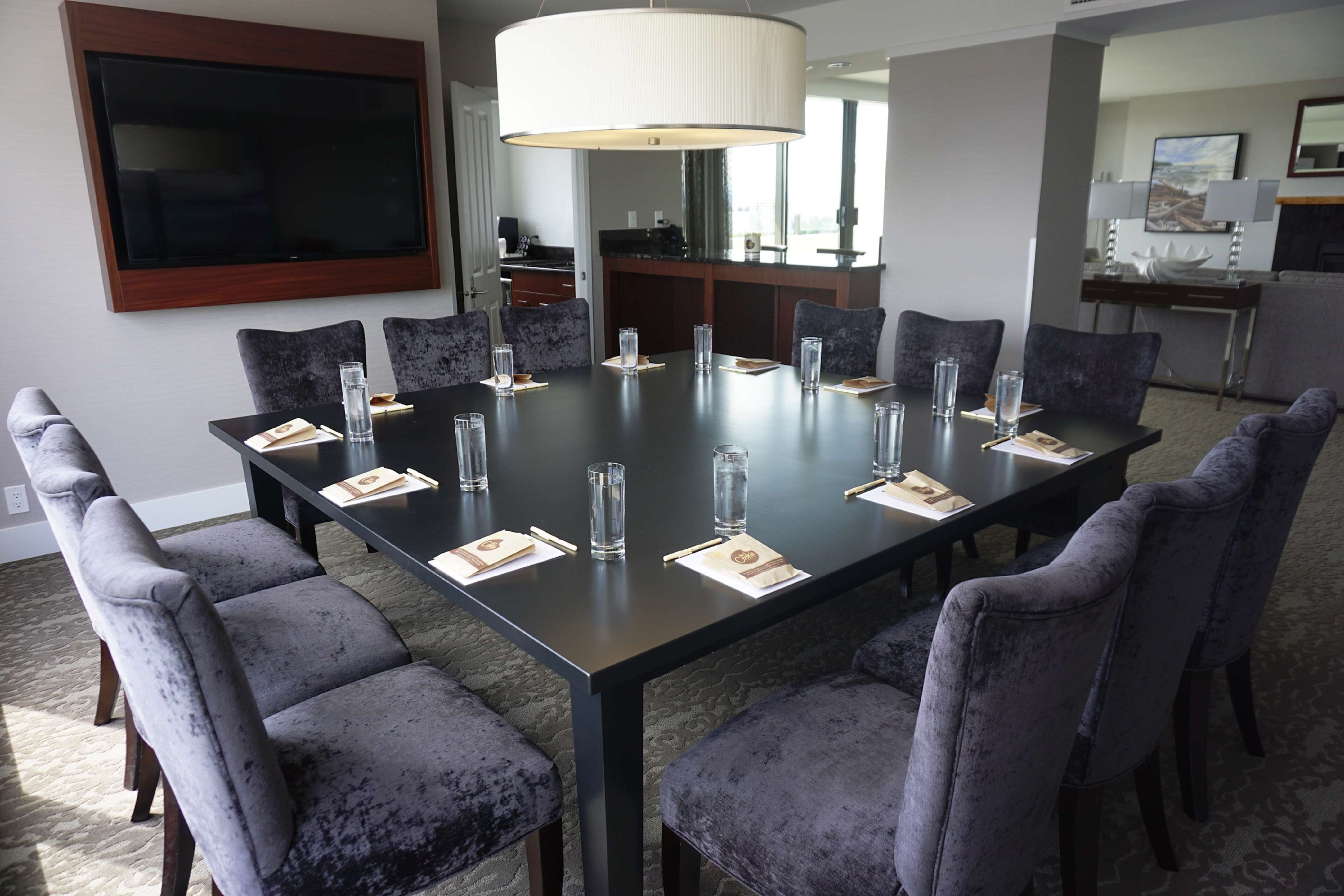 Une salle de conférence avec une grande table, des chaises, un téléviseur et un coin cuisine à l’arrière-plan.