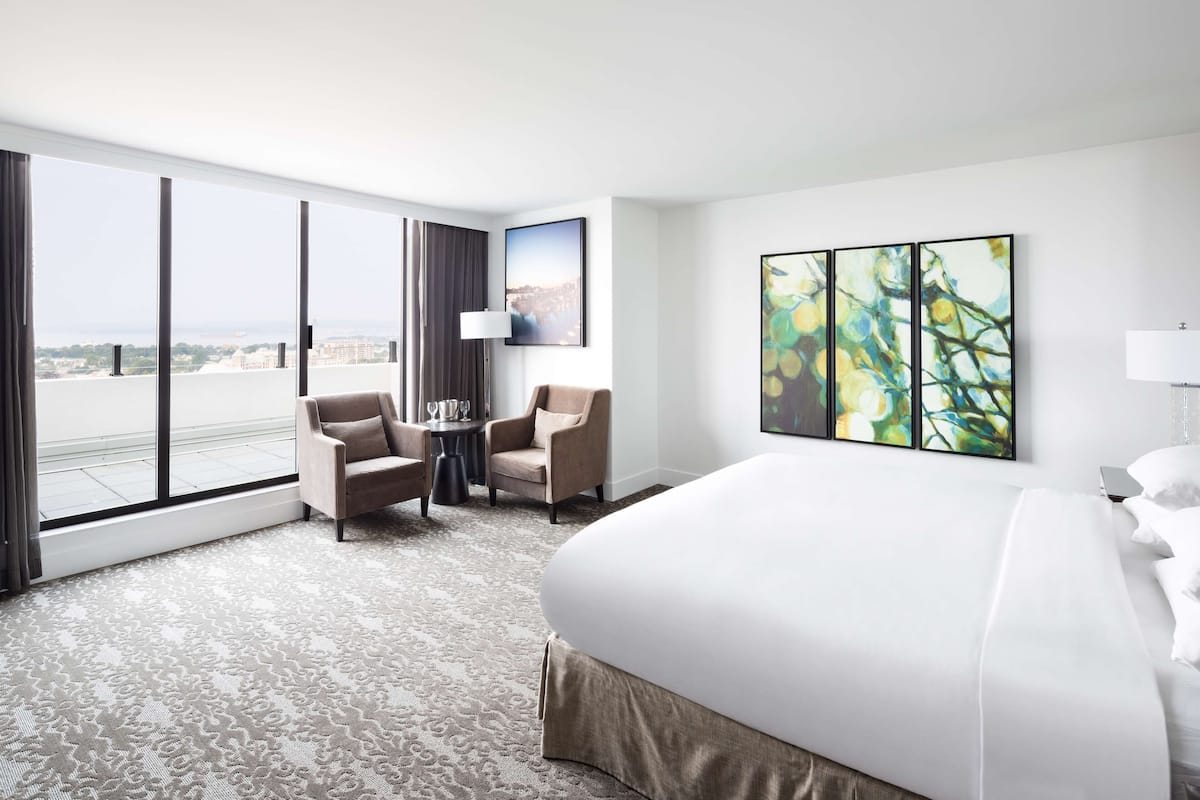 Habitación de hotel con una cama grande, dos sillones, una mesita y vistas a la ciudad a través de ventanales del suelo al techo.