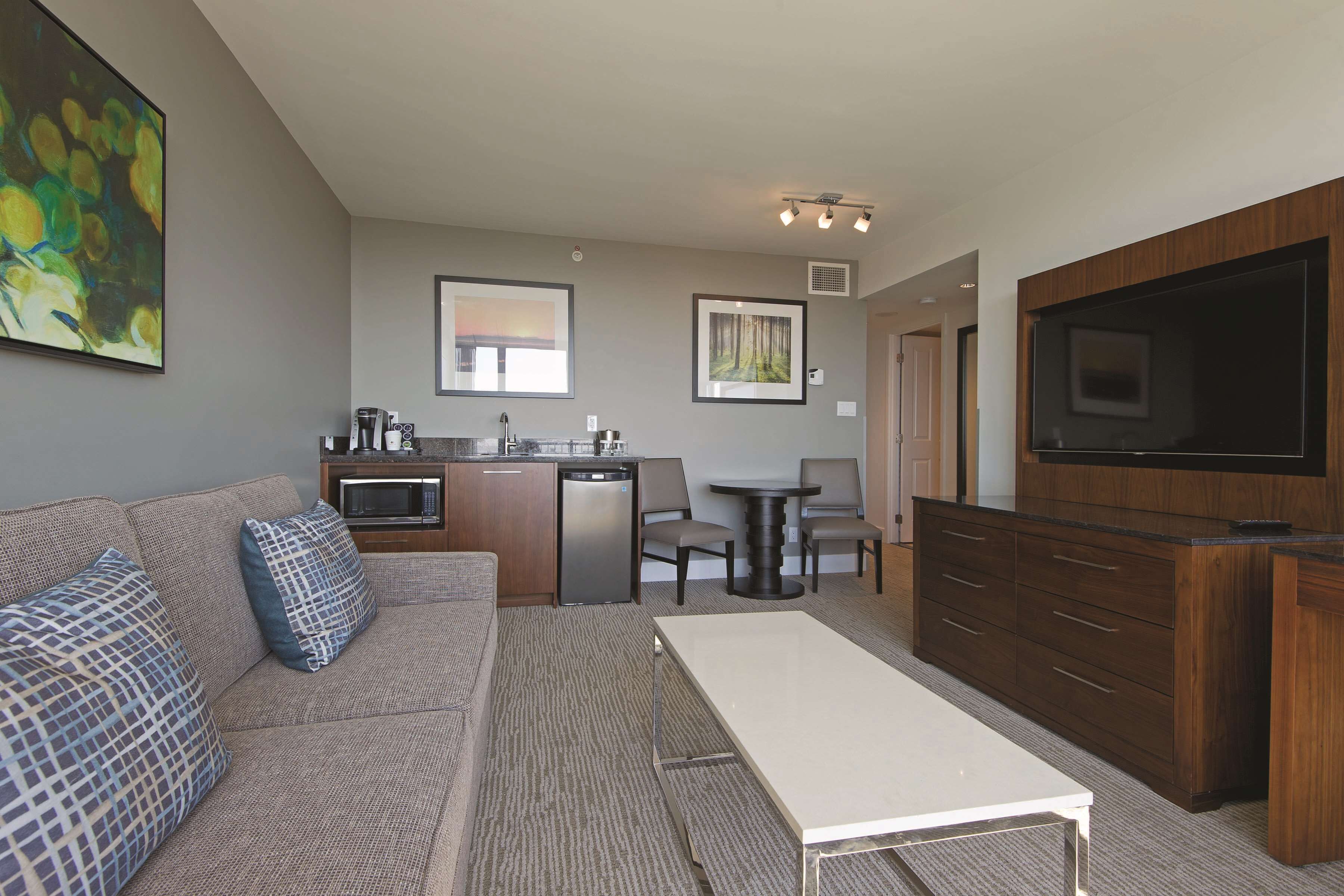 A compact living space with a kitchenette, a sofa with patterned cushions, a dining table, and a flat-screen TV mounted on a wooden cabinet.