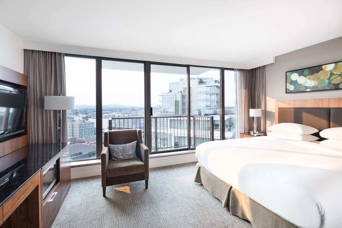 Habitación de hotel con una cama grande, televisión, una silla y vistas de la ciudad a través de amplias ventanas.