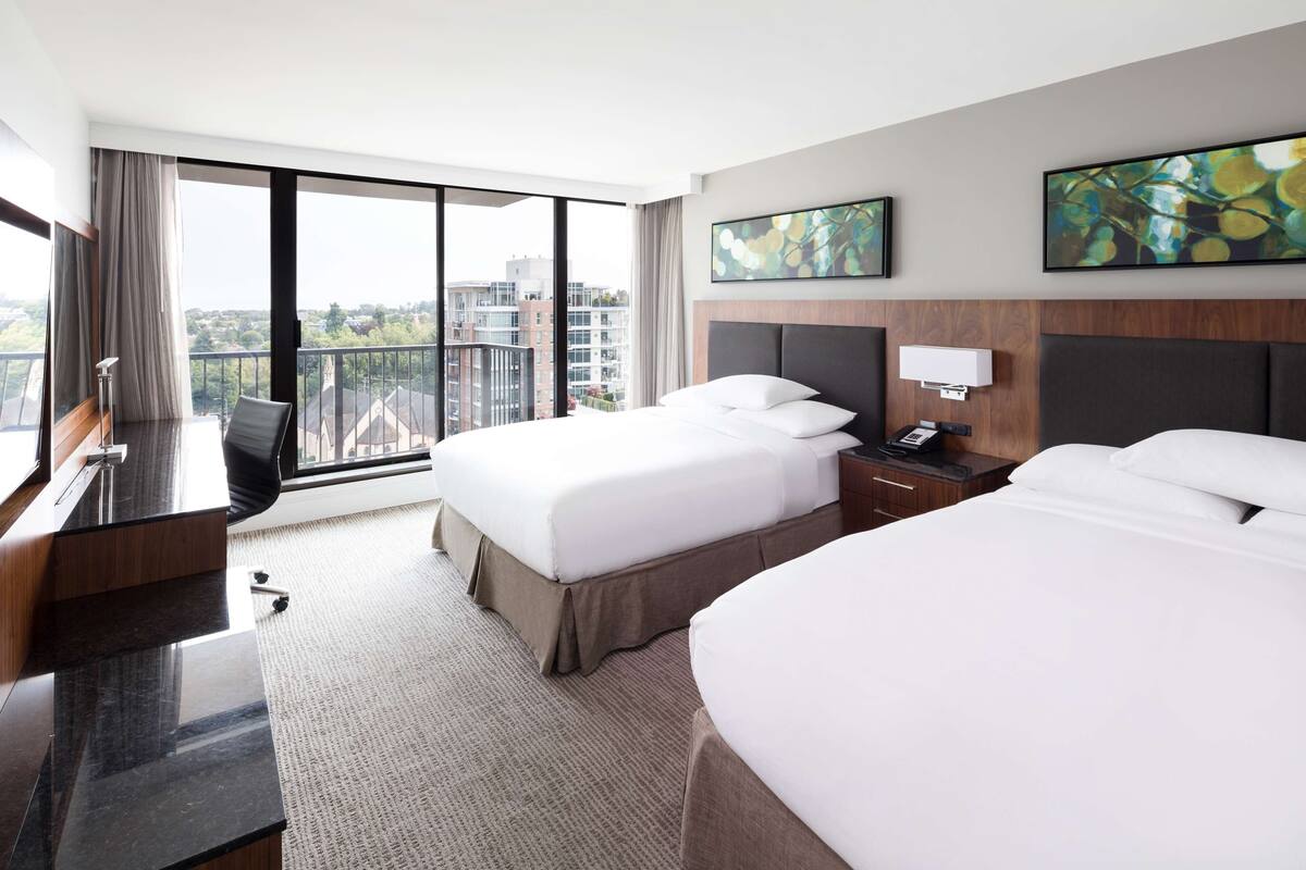 Habitación de hotel con dos camas, un escritorio y un amplio ventanal con vistas a la ciudad.