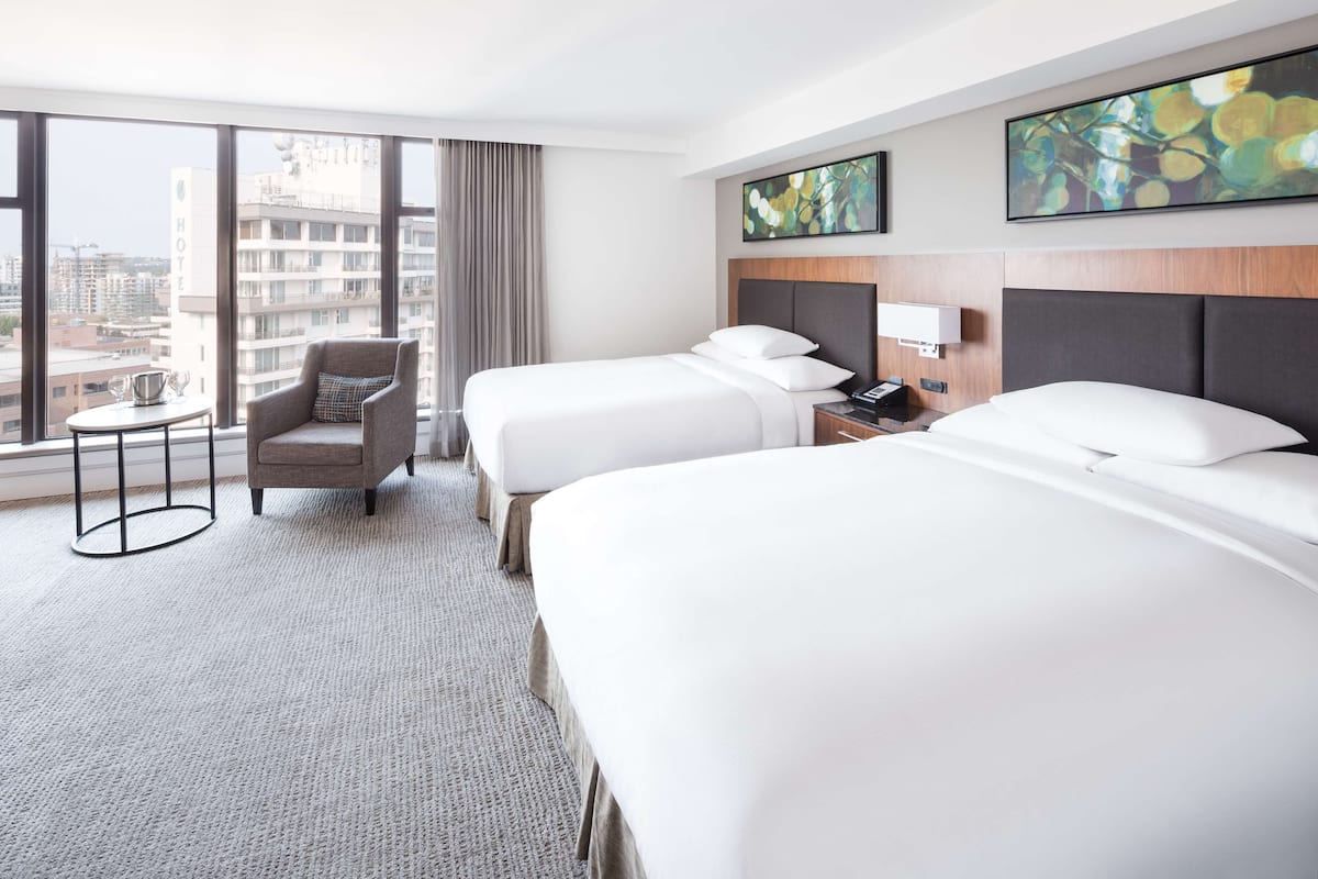 Habitación de hotel con dos camas, una silla, una mesita y vistas a la ciudad.